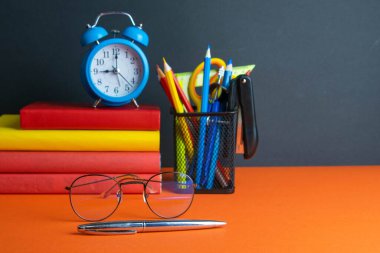 Bir yığın kitap, ön planda kadın gözlüğü, mavi saat ve turuncu arka planda duran bir bardak kalem..