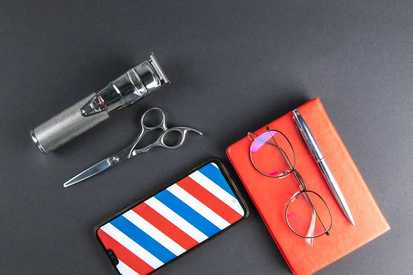 A machine for haircuts, scissors, comb stand on a white towel and next to there is a Phone, pen and a red notebook on which there are women\'s glasses.