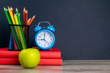 Mavi çalar saat, çok renkli kalemlerle birlikte kırmızı kitapların üzerinde duruyor, bir elmanın yanında..
