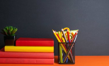 Bir yığın kitap, yeşil bir çiçek ve turuncu arka planda duran bir bardak kalem..