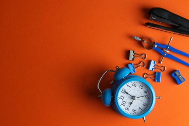 Kalemler, mavi alarm saati, pusula, cetvel ve zımba turuncu arka planda..