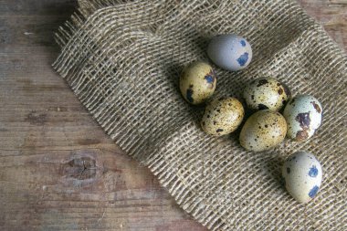 Bıldırcın yumurtaları bir çiçeğin yanındaki tahta masadaki çuval bezi üzerinde duruyor. Açık alanlı en üstteki görünüm.