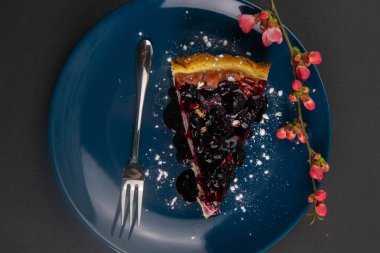 Kırmızı çiçekli mavi bir tabakta, aynalı dutlu yaban mersinli kek dilimleri ve nane yaprakları siyah masanın üzerindedir. Üst Görünüm.