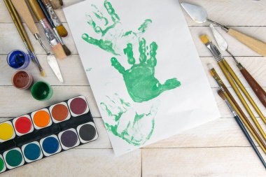Sanatçı aksesuar koleksiyonu. Tuval, boya tüpü, sanat fırçaları, palet bıçağı ve ahşap masada duran yeşil boyalı çocukların el izleri olan bir mektup. Sanatçı atölyesi arka planı.
