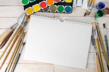 Sanatçı aksesuar koleksiyonu. Tablo, boya tüpü, sanat fırçaları palet bıçağı ve ahşap masanın üzerinde duran beyaz defter. Sanatçı atölyesi arka planı.