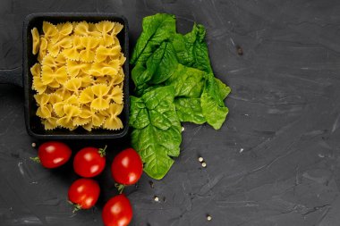 Kızartma tavasında çiğ makarna, domates ve arka planda ıspanak. Açık alanlı en üstteki görünüm.