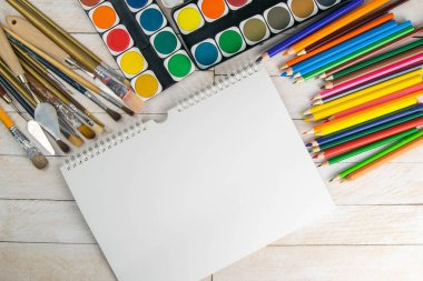 Sanatçı aksesuar koleksiyonu. Tablo, boya tüpü, sanat fırçaları palet bıçağı ve ahşap masanın üzerinde duran beyaz defter. Sanatçı atölyesi arka planı.