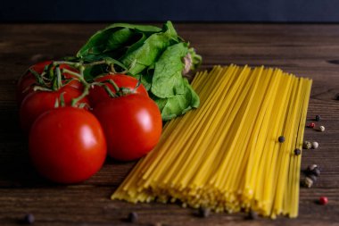 Sebze makarna malzemeleri: spagetti, biber, domates, fesleğen, biberiye, zeytin yağı, sarımsak, deniz tuzu ve koyu ahşap zemin üzerindeki baharatlar. Ön manzara, yakın plan..