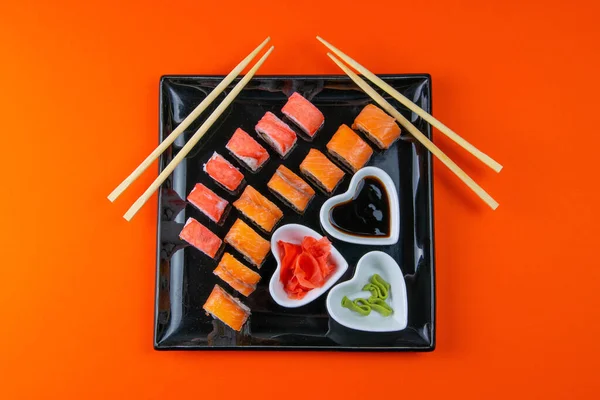 Black plate with sushi, filled with rolls of different kinds, next to stand sticks for sushi, red marinaginger and soy sauce on a orange background. Top views