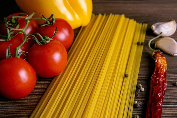 Vegetable pasta ingredients: spaghetti, peppers, tomatoes, basil, rosemary, olive oil, garlic, sea salt and spices on a dark wooden background. Front views, close-up.