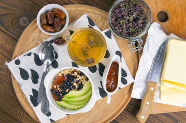 Yanında yoğurt, hurma ve kayısı reçeli bulunan yulaf ezmeli, yabanmersinli ve avokadolu sağlıklı bir kahvaltı. Üst Görünüm.