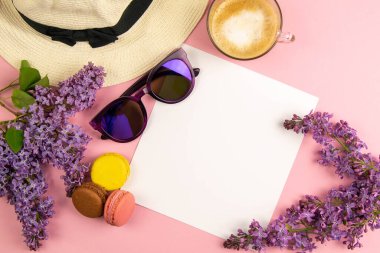 Etrafı leylak renkleri, güneş gözlükleri, renkli badem kurabiyeleri ve plaj şapkasıyla çevrili boş bir çarşaf. Şık, minimalist iş yeri konsepti. Kopyalama alanı olan üst görünüm.