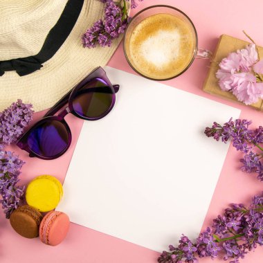 Etrafı leylak renkleri, güneş gözlükleri, renkli badem kurabiyeleri ve plaj şapkasıyla çevrili boş bir çarşaf. Şık, minimalist iş yeri konsepti. Kopyalama alanı olan üst görünüm.