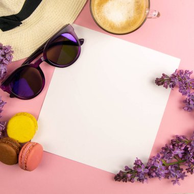 Etrafı leylak renkleri, güneş gözlükleri, renkli badem kurabiyeleri ve plaj şapkasıyla çevrili boş bir çarşaf. Şık, minimalist iş yeri konsepti. Kopyalama alanı olan üst görünüm.