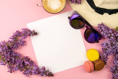 Etrafı leylak renkleri, güneş gözlükleri, renkli badem kurabiyeleri ve plaj şapkasıyla çevrili boş bir çarşaf. Şık, minimalist iş yeri konsepti. Kopyalama alanı olan üst görünüm.