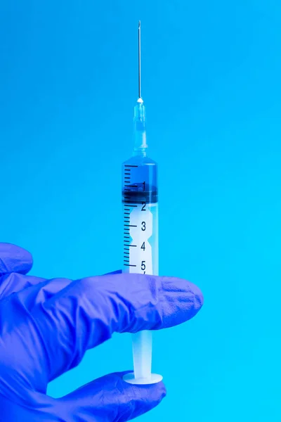 A hand in a blue glove holds a syringe. Doctor holds a syringe for vaccination.Coronavirus. Medicine. Blue background, close-up.