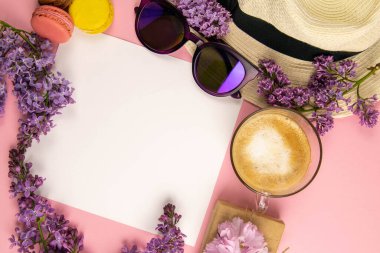 Etrafı leylak renkleri, güneş gözlükleri, renkli badem kurabiyeleri ve plaj şapkasıyla çevrili boş bir çarşaf. Şık, minimalist iş yeri konsepti. Kopyalama alanı olan üst görünüm.