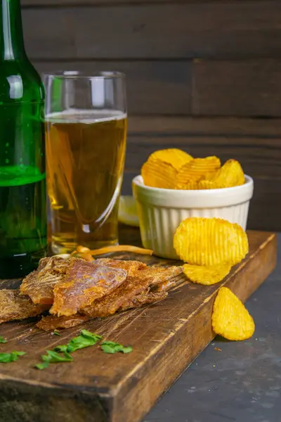 stock image Dried cod with pepper with beer, lemon and potato chips on dark wooden board. Snack on fish with beer. Close-up.