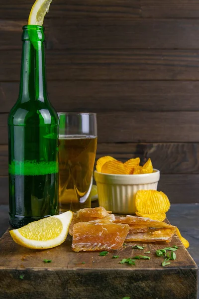 Dried cod fillet with beer, lemon and potato chips on dark wooden board. Snack on fish with beer. Front views, close-up.