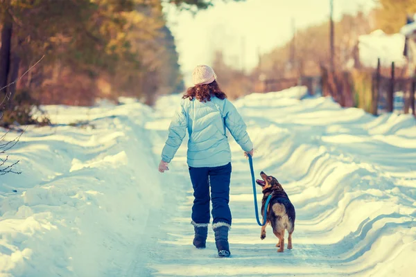 年轻的女人与她的狗在村里的雪路上走回相机 — 图库照片