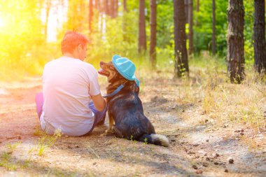 Bir adam ormandaki yolda bir köpekle sırtını kameraya ile oturuyor. Köpek güneş şapka giyiyor
