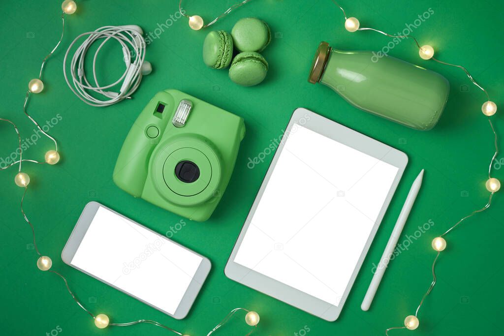 Flat lay tablet, phone, analog camera and headphones, bottle and macaroons on green background 