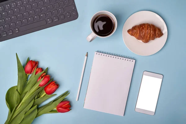 Taza de café, croissant, bloc de notas, pluma, teléfono, teclado y flores — Foto de Stock
