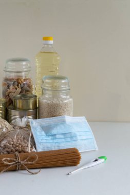 Karantina sırasında uzun süre depolanan çeşitli ürünler. Makarna, yulaf ezmesi, pirinç, nohut, kahve, konserve sebze, maske ve termometreyle birlikte şişedeki yağ. Coronavirus konsepti - evde kal, güvende ol.