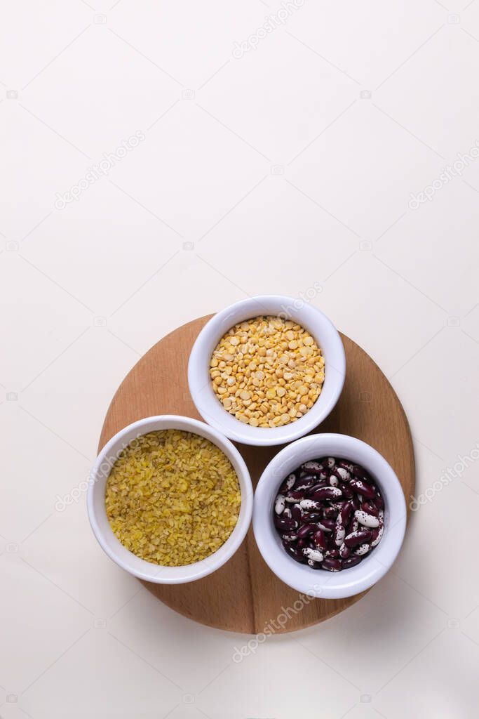 Bulgur, beans, peas in white bowls on a wooden cutting board on a light yellow background. Healthy food concept. Vertical orientation. Top view. Copy space.
