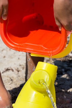 Çocuk, kırmızı kovasından kumsaldaki sarı sulama kutusuna deniz suyu döküyor.