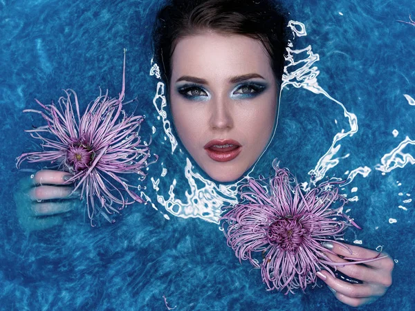 Menina modelo de moda bonita tomando banho azul, spa e conceito de cuidados com a pele. Jovem com maquiagem brilhante e flores rosa relaxando em banho azul. Rosto e mãos saudáveis . — Fotografia de Stock