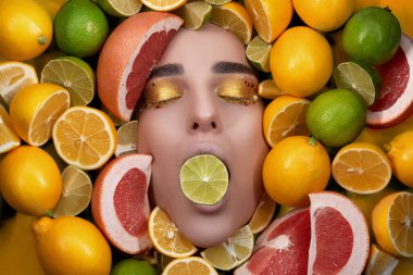 Parlak makyajlı güzel bir manken ağzında limon, limon ve turunçgilin arka planında limon tutuyor. Üst Düzey Erişim