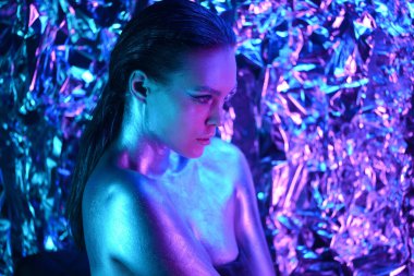 Uzun saçlı, yuvarlak güneş gözlüklü, siyah parlak elbiseli moda kızı stüdyoda neon ışıkta poz veriyor.