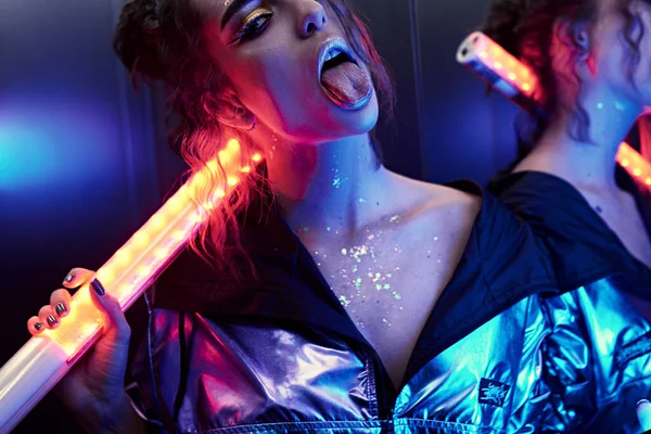 Mulher bonita com brilhos em seu rosto em luz ultravioleta mostrando sua língua com cara boba. Arte colorida compõem — Fotografia de Stock