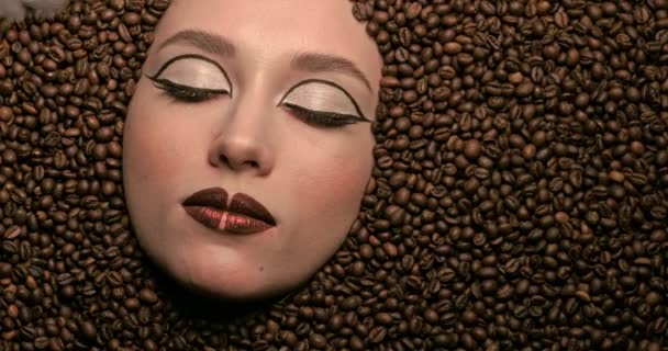 Aromatyczne ziarna kawy wokół twarzy pięknej młodej kobiety i lekkiego dymu. Styl życia kawy. ładna dziewczyna blokująca przed aparatem piękny brązowy makijaż. Portret z bliska. Wysokiej jakości 4k wideo. — Wideo stockowe