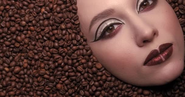 Close-up portret van een high fashion model vrouw ondergedompeld in koffiebonen met een lepel in haar mond. Mooie jonge meisje schoonheid make-up. Ik kijk naar de camera. Koffie levensstijl. Hoge kwaliteit 4k video — Stockvideo