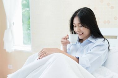 Asyalı genç kadın bedroo yatakta otururken kahve içme