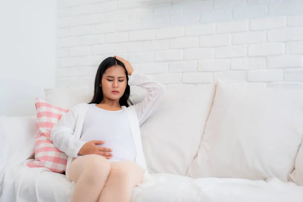 Молода вагітна жінка з головним болем сидить на дивані в живому роу — стокове фото