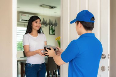 Asyalı teslimatçı mavi üniformalı genç adam evin önünde yemek kutuları tutuyor ve Asyalı kadın teslimatçıdan gelen yiyecek kutularını kabul ediyor. Reklamcılık, İş, Ulaşım Konsepti.