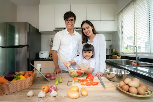 Asian family with father, mother and daughter shredded vegetable salad and look at camera while the family was cooking in the kitchen at home. Family life love relationship, or home fun leisure activity concep