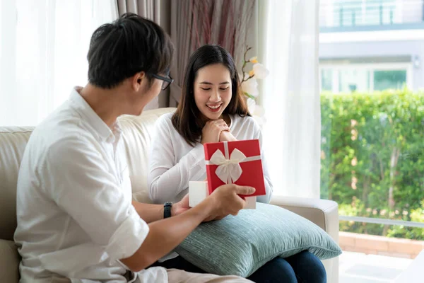 Asian man give a woman a red gift box. She look at the gift in the box and surprise at the wedding anniversary or her birthday in living room at house. Love, relationship, wedding, or relaxing casual lifestyle concept