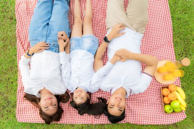 Asian teen family happy holiday picnic moment in the park with father, mother and daughter lying on mat and smile to happy spend vacation time togerter in green garden with friut and food
