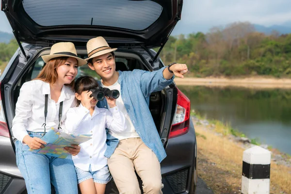 Portrait of Asian family sitting in car with father pointing to view and mother holding maps with daughter looking beautiful landscape through binoculars while vacation together in holiday. Happy family time.