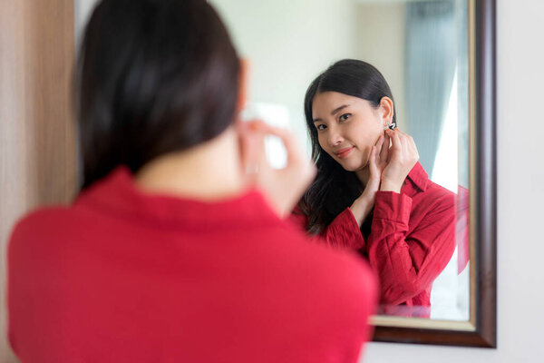 Beautiful Asian Woman wearing red dressed putting star earring looking in mirror in her bedroom at home. Makeup in morning getting ready before going to work.