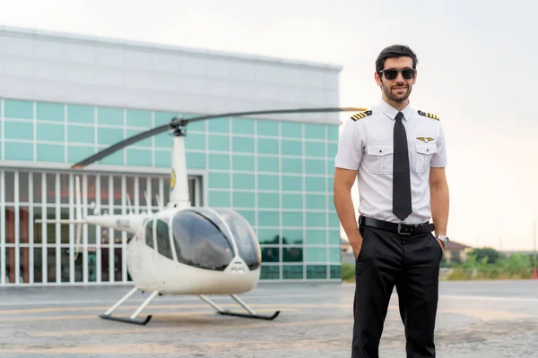 英俊潇洒的商业飞行员身穿白色机长制服 站在一架小型私人直升机旁边 背景是国际机场的一个配备私人喷气式候机楼的着陆点 看上去很聪明 — 图库照片