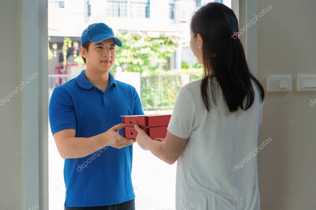 Asian delivery young man in blue uniform smile and holding pizza boxes in front house and Asian woman accepting a delivery of pizza boxes from deliveryman. Advertising, Business, Transportation Concept.