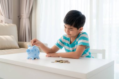 Küçük Asyalı çocuk evdeki oturma odasındaki beyaz bankaya para koyuyor. İşletme geleceği için yatırım yapıyor.