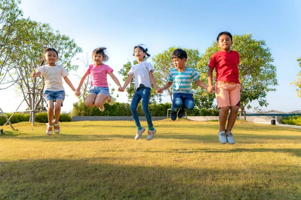 Large group of happy Asian smiling kindergarten kids friends holding hands playing and jumping together during a sunny day in casual clothes at city park.