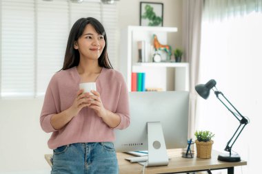 Asyalı güzel genç bir kadın evde bilgisayar başında çalışıyor ve kahvesini içerken evinde oturma odasında dinleniyor. Evdeki sosyal mesafeli yaşam tarzı