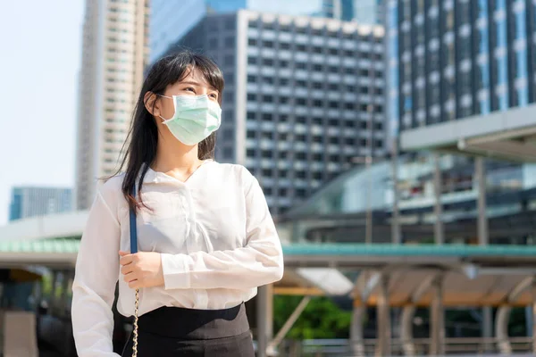Young stress Asian businesswoman in white shirt going to work in pollution city she wears protection mask prevent PM2.5 dust, smog, air pollution and COVID-19 with business office building in Bangkok, Thailand.